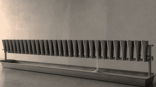 Lange Metallhalterung mit rund dreissig gleichartigen Blechtrichtern ueber einer gemeinsamen Sammelschale, aus der nur ein einziger Trichter Sand rieseln laesst
