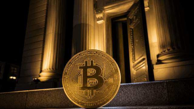Goldene Muenze mit eingepraegtem Bitcoin-Zeichen auf der Treppenstufe vor dem beleuchteten Saeulenportal eines alten Bankgebaeudes bei Nacht