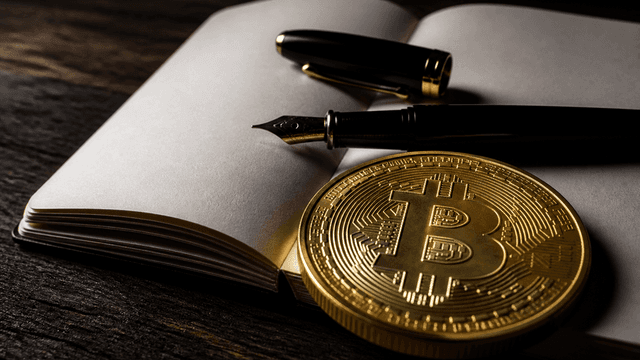 Aufgeschlagenes leeres Formularheft aus Buettenpapier auf dunkler Eichenholzplatte, daneben ein schwarzer Fuellfederhalter mit offener Feder, davor eine schwere Goldmuenze mit eingepraegtem Bitcoin-Zeichen